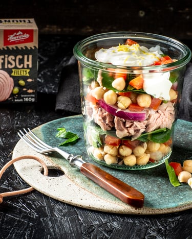 In einem Glas sind Kichererbsen, Thunfisch, Tomate, Spinat und Zwiebel mit einem Klecks Dressing on top übereinander geschichtet.