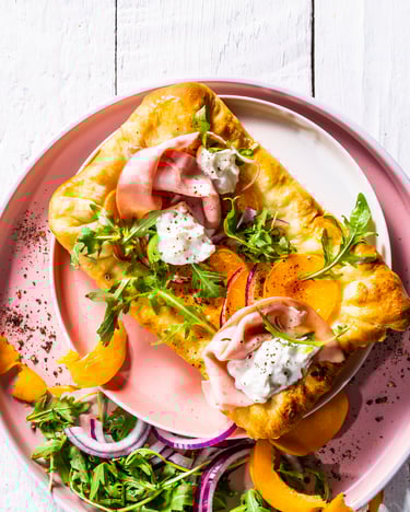Rechteckige Mini-Pizza liegt auf einem rosa Teller, belegt mit Mortadella, Burrata, Aprikosen und Rucola