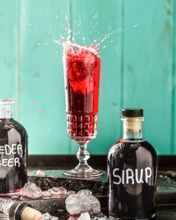 Aus einem hohen Glas spritzt Fliederbeersirup heraus. Davor stehen zwei selbstbeschriftete Flaschen gefüllt mit rotem Sirup.
