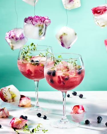 In zwei Weingläsern Blueberry Gin Spritz mit Eiswürfeln, und Blaubeeren. Um Die Drinks herum und in der Luft hängen Eiswürfel mit Blüten.
