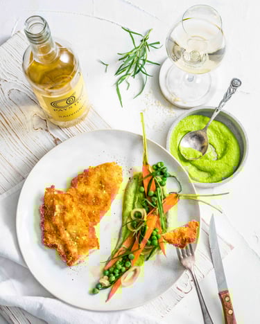 Ein Teller mit zwei panierten Hähnchenschnitzeln neben Erbsen und Möhren und daneben eine Schale mit cremigem Erbsenpüree auf einem hellen Holzuntergrund neben einem Glas und einer Flasche Sauvignon Blanc