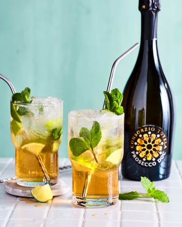 Zwei Gläser mit Strohhalm und gelb-orangem Projito, der mit frischen Minzeblättern dekoriert ist, auf einer weißen Fliesenfläche vor türkisem Hintergrund. Daneben steht eine Flasche Prosecco DOC.