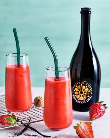 Zwei Cocktailgläser mit eisgekühltem Erdbeer Prosecco Slush auf einem hellen Untergrund neben frischen Erdbeeren und Vanilleschoten vor türkisem Hintergrund. Dazu eine Flasche Prosecco DOC.
