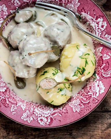 Königsberger Klopse mit Kartoffeln und Kapernäpfeln in einem Suppenteller auf dunklem Holzuntergrund.