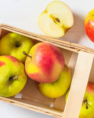 Grün-rote Äpfel ganz und halbiert in hellem Holzkörbchen
