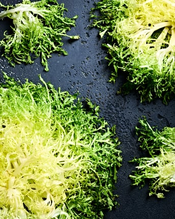 Frisée-Salat als ganzer und halber Kopf mit sichtbar heller Mitte auf einem dunklen Untergrund