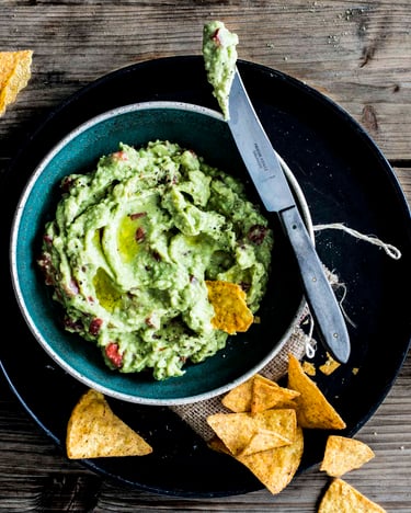 Guacamole auf klassische Art in einer Schüssel, drum herum liegen Nachochips zum Dippen, ein Messer mit Guacamole auf der Messerspitze liegt am Schüsselrand. Fotografiert auf hölzernem Untergrund von oben.