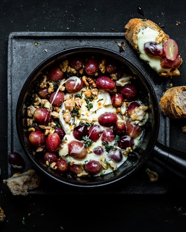 Eine Pfanne mit Baked Brie und Trauben aus dem Ofen vor dunklem Hintergrund mit zwei gerösteten Brotscheiben zum Dippen