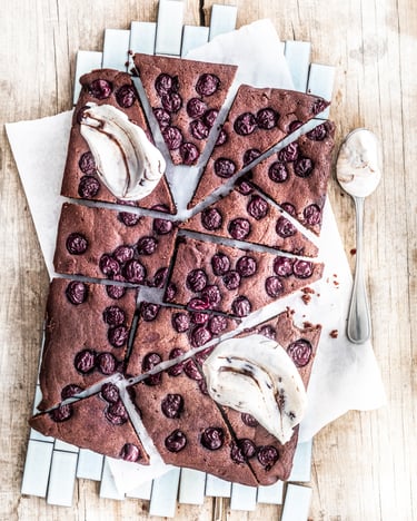 Vegane Schoko-Kirsch-Brownies mit Nugat-Kokossahne und einem Esslöffel voll mit Sahne auf einem hellen Holzuntergrund.