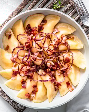 Im Kreis aufgefächert sind die Pieroggen in einer Pfanne drapiert und mit Zwiebeln und Speck dekoriert.