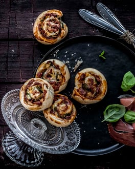 Blätterteigschnecken auf einer Servierplatte und einem Teller neben Parmaschinken und Basilikum und Besteck