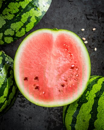 Ganze und angeschnittene Wassermelonen mit rosarotem Fruchtfleisch und wenig Kernen als Topshot vor dunkelgrauem Untergrund