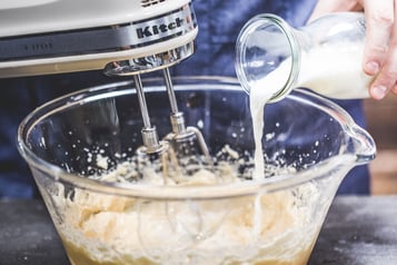 In einer großen Glasschüssel mit hellem Rührteig rührt ein Handrührgerät. Eine Hand kippt aus einer Glaskaraffe Milch hinzu.