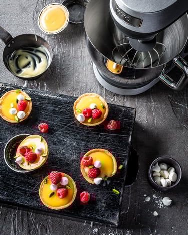 Mehrere Zitronen-Himbeer-Tartelettes auf schwarzem Brett und Krups Standmixer.