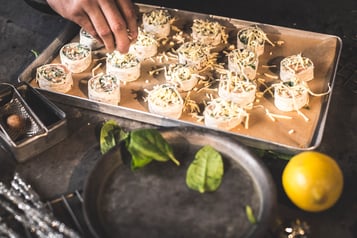 Tortillaschnecken sind auf einem Backblech aufgereiht und werden von einer Hand mit Käse bestreut