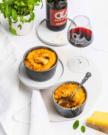 Zwei Formen mit Shepherd's Pie it Kürbispüree stehen mittig auf weißem Untergrund. Im Hintergrund eine Flasche Rotwein, daneben ein befülltes Weinglas.