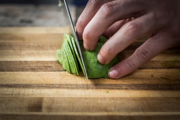 Eine halbe Avocado wird auf einem Holzbrett mit einem Messer  in feine Streifen geschnitten