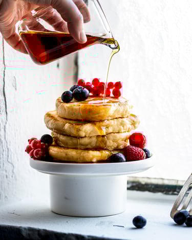 Fluffige Pancakes sind auf einem Podest aus Schüssel und Teller übereinandergestapelt und dekoriert mit gemischten Beeren. Gerade wird Ahornsirup aus einer Glaskaraffe darüber gegossen.