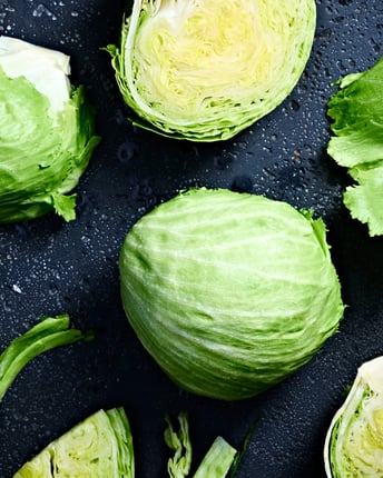 Ein halbierter und ein ganzer Eisbergsalat neben losen Eisbergsalatblättern auf einem dunklen Untergrund
