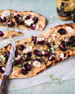 Flammkuchen mit Brombeeren und Thymian auf weißem Backpapier neben einem Senfglas.
