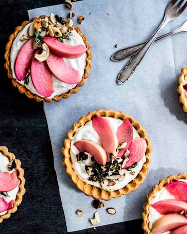 Selbst gebackene Mini-Tartelettes mit Pfirsich, Joghurtcreme und Nusscrumble auf Backpapier mit Kuchengabeln.