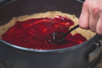 Mit einem Teelöffel wird Himbeermarmelade auf dem Teig in der Form verteilt.