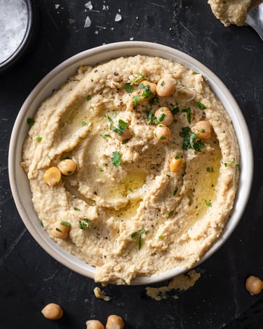 Eine große Schale mit Hummus, bedeckt mit Olivenöl, Kräutern und Kichererbsen, ist von oben fotografiert. Daneben liegen Kichererbsen verteilt und eine kleine Schale mit Meersalz.