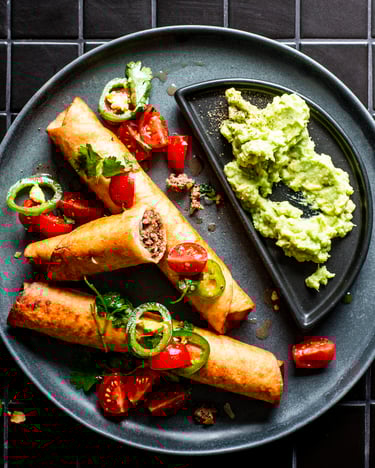 Zwei ganze und eine angeschnittene Beef Flautas mit Hackfleischfüllung, Salsa-Topping und Guacamole als Dip auf einem dunklen Teller über einem schwarzen Fliesenuntergrund