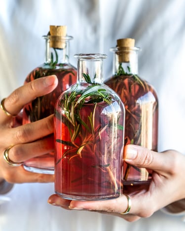 Person in weißem Hemd hält 3 Flaschen mit Korken, pinkem Sirup und Rosmarin in der Hand.