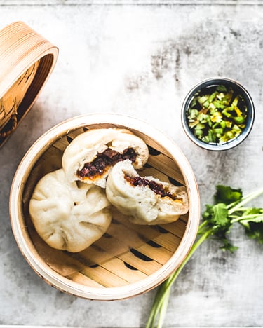 Ein ganzer und einhalbierter Dumpling, bei dem die Schweinebauch-Füllung sichtbar ist, liegen zusammen in einem Bambus-Dampfkorb neben frischer Petersilie und dem angelehnten Bambusdeckel