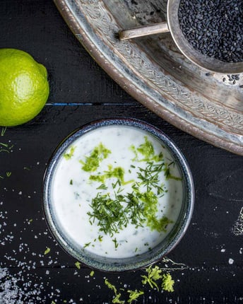 Joghurt-Limetten-Dressing in silberner Schale angerichtet, fotografiert von oben auf dunklem Untergrund. Daneben Limetten und -abrieb, eine Tasse voll dunklem Sesam steht ebenfalls im Bild.