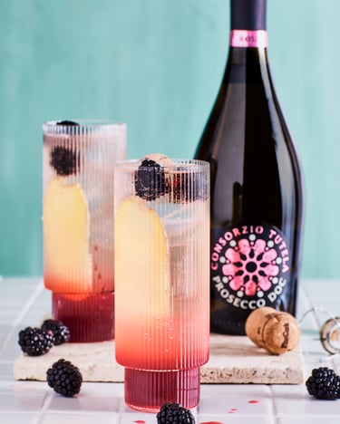 Zwei Gläser mit Brombeer Mule mit Prosecco Rosé. An der Glasseite ist eine längs geschnittene Ingwerscheibe zu sehen, oben schwimmen Eiswürfel und frische Brombeeren sind in Glas und auf dem weißen Fliesenuntergrund verteilt. Daneben steht eine Flasche Prosecco DOC Rosé