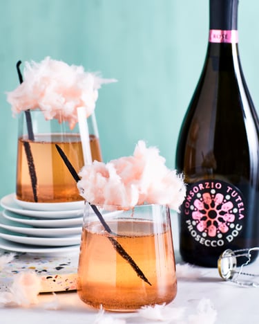 Zwei Gläser mit Prosecco Rosé, die mit hellrosa Vanille-Zuckerwatte und einer Vanilleschote getoppt sind, vor weißem Untergrund und einer türkisen Wand. Daneben eine Flasche Prosecco DOC Rosé.