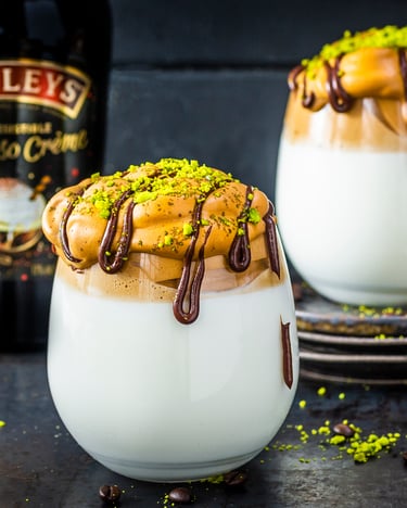 Zwei Gläser, die üppig gefüllt sind mit Milch und Dalgona Creme stehen auf einer dunklen Tischplatte. Dekoriert ist die Creme mit Schokoladensoße und gemahlenen Pistazien.