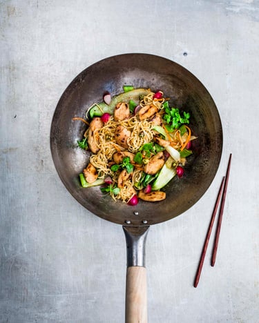 Wok mit dünnen Nudeln, Hähnchen und Pak Choi auf einem Betonuntergrund. Daneben zwei Essstäbchen