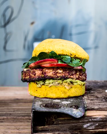 Ein prall gefüllter Burger mit gelb-orangenem Möhren-Bunist neben dem Kidneybohnen-Patty mit Avocado, Nektarinen und Babyspinat gefüllt.