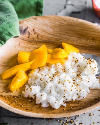 Sticky Rice mit Kokosmilch und Mango in einem Bambusteller mit einem Löffel und einem grünen Tuch nebenliegend.