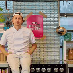 Hannes sitzt auf einem Herd. Im Hintergrund hängt ein Poster auf dem "Eine Flocke Butter" steht.