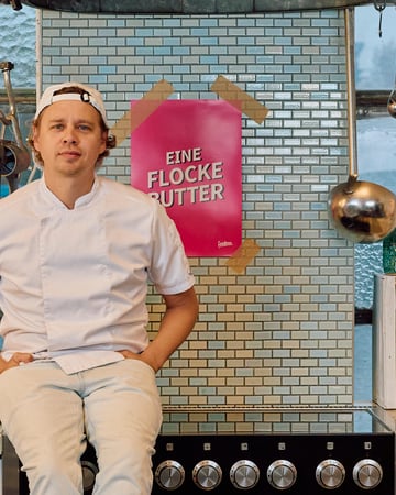 Hannes sitzt auf einem Herd. Im Hintergrund hängt ein Poster auf dem "Eine Flocke Butter" steht.