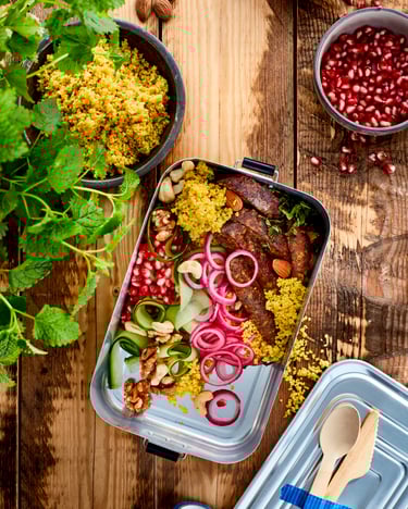 Couscous mit Jackfruit-Streifen, roten Zwiebelringen, Gurkenstreifen, Granatapfelkernen und Nüssen in silberner Brotdose auf Holzuntergrund