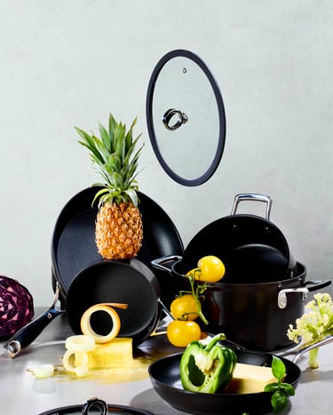 schwarze Töpfe und Pfannen mit Deckeln angerichtet, umgeben von buntem Obst und Gemüse