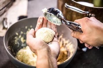 Teig für Quarkbällchen wird mit einer Hand und einem Eisportionierer in Bällchen geformt. Im Hintergrund eine große graue Schüssel mit Teig.