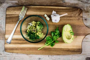 Auf einemHolzbrett liegen Avocado und Kräuter, daneben steht eine kleine Glasschüssel, daneben liegt eine Gabel