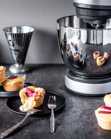 Schwarzer Tisch mit Krups Standmixer, schwarzen Tellern, Gabeln, Messbecher und mehreren Rhabarber-Brioche-Toertchen mit Vanillecreme.