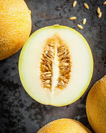 Eine halbierte Netzmelone der Sorte Galia neben ganzen Melonen auf dunkelgrauem Untergrund