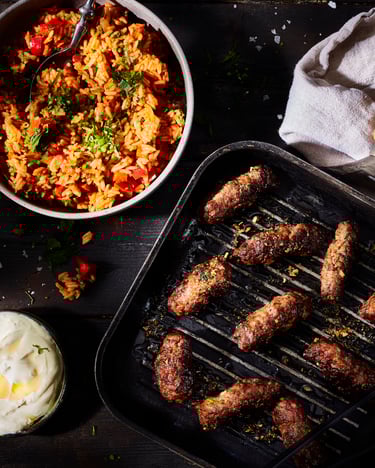 Cevapcici in einer Grillpfanne, daneben steht eine Schale mit Djuvec-Reis.