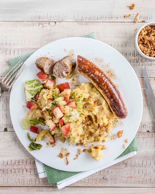 Bratwurst mit Kartoffelbrei und Apfel-Wirsing auf einem Teller serviert nebenliegend Besteck.