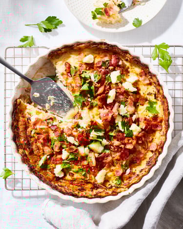 Eine runde Auflaufform mit einer angeschnittenen Quiche mit einem Boden aus Kartoffelpuffer und einem Topping aus Petersilie, Bacon und Pecorino, vor einem hellen Untergrund