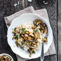 Auf einem weißen Teller, der auf einem grauen Tuch steht ein Risotto mit Steinpilzen und Kräutern. Darin liegt ein Esslöffel. Oben im Bild ein Stück Parmesan