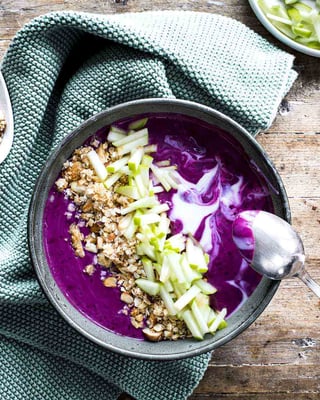 Lila Heidelbeer-Smoothie-Bowl mit Topping aus Apfelstücken und Granola auf Holzgrund und helltürkisem Tuch
